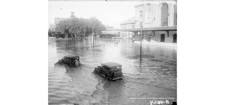 1933 Union Station surrounded by flood water with two cars driving through the water.