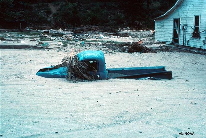 A blue pickup truck is submerged up to its hood in floodwater during the 1976 flood of the Big Thompson River
