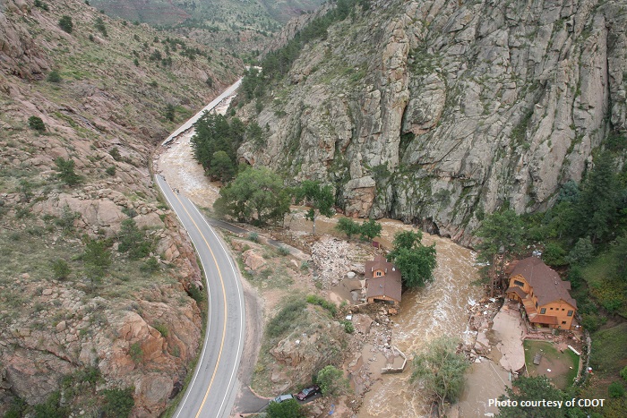 A flooded river cutting between two houses and across a washed out road in a mountain canyon