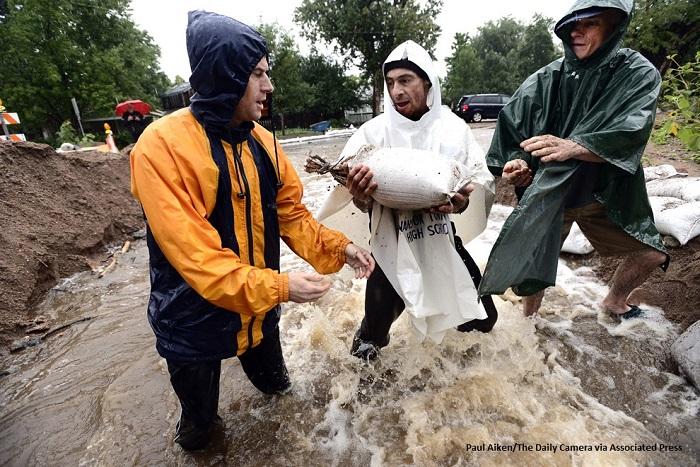 Three men in rain coats pass along a sandbag while standing in rushing floodwaters