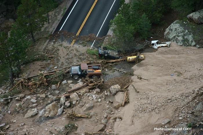 Several vehicles lie partially buried in mud and debris at the base of a mudslide that covers a two lane mountain road.