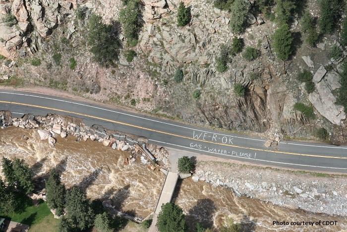Overhead view of a partially washed out two lane road in a mountain canyon with a message written on the road reading: We R OK Gas Water Please!