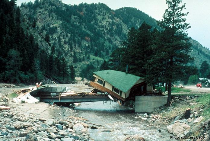 A ruined green-roofed cabin resting precariously on a bridge across the Big Thompson River with mountains in the background in 1976